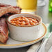 A plate of ribs and beans with a vanilla bean stoneware ramekin of sauce on a table.