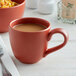 A red Libbey Driftstone mug filled with coffee on a table with a bowl of cereal and a spoon.