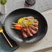 An Acopa matte black stoneware coupe plate with steak and carrots on it.