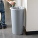 A person standing next to a Rubbermaid Untouchable gray round wastebasket and putting a paper cup inside.