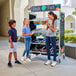 A woman and kids standing next to a Cambro Fresh & Fast fruit stand with grey and blue merchandiser graphics.