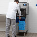 A man in a white shirt and gray pants standing next to a Manitowoc ice machine with blue ice bin.