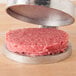 A raw hamburger patty being pressed on a metal plate with the American Metalcraft 4 1/2" Adjustable Cast Aluminum Hamburger Press.