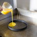 A person pouring yellow liquid into a pan using a Prince Castle omelet ring with a black handle.