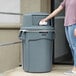 A woman using a white towel to open a Rubbermaid BRUTE trash can.