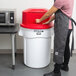 A person wearing an apron putting a red Rubbermaid BRUTE trash can in a kitchen.
