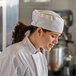A woman in a white chef's uniform wearing a white mesh top chef skull cap.