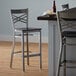 A Lancaster Table & Seating cross back bar stool with a black wood seat next to a table.