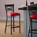 A black Lancaster Table & Seating cross back bar stool with a red vinyl padded seat next to a table.