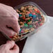 A hand holding a clear plastic Dart deli container filled with nuts and candy.