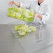 A woman in a white chef's coat holding a clear Cambro food pan with lettuce on a tray.