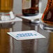 A glass of beer next to a white square paper coaster with a blurry hand in the background.