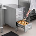 A chef using tongs to put food in a ServIt narrow freestanding drawer warmer.