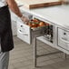 A person in a black apron opening a silver ServIt built-in drawer warmer with food inside.