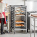 A woman in a red apron standing in a school kitchen with a Cambro sheet pan rack full of pastries.