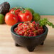 A charcoal Perfecto Molcajete filled with salsa with tomatoes and green peppers on a table.