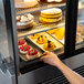 A hand reaching into a Vollrath glass display case to grab a slice of cake.