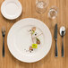 A RAK Porcelain ivory wide rim porcelain flat plate with food and silverware on a wooden surface.