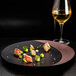 A RAK Porcelain bronze and black flat plate with food and a glass of wine on a black surface.