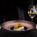 A RAK Porcelain bronze and black gourmet deep plate with food on a table.