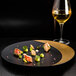 A RAK Porcelain gold and black deep plate with food and a glass of yellow liquid on a black surface.