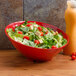 A red slanted bowl filled with salad next to a bottle of juice.