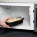 A hand placing a Pactiv Newspring VERSAtainer container of food in a microwave.