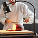 A man in a white coat using an Avantco stainless steel heat lamp to cut a piece of food on a table.