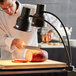 A man using an Avantco black dual arm heat lamp to cut meat on a cutting board.