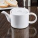 An Arcoroc white teapot with a lid on a table.
