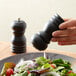 A hand holding a black Acopa pepper mill over a salad.