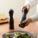 A person using an Acopa matte black wooden pepper mill to add pepper to a plate of salad.