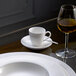 A white Villeroy & Boch saucer with a white cup on it and a glass of wine on a table.