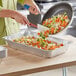A pan filled with broccoli, carrots, and red vegetables being poured into a Choice Full Size Foil Steam Table Pan.