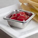 A small stainless steel square sauce cup filled with ketchup on a table with french fries.