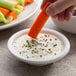 A person holding a carrot dipping it into white sauce.