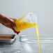 A hand holding a Tablecraft 3-sided silicone measuring cup pouring yellow liquid into a glass bowl.
