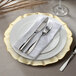 A white Charge It by Jay glass charger plate with a gold rim set on a white surface with silverware and a white napkin.