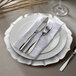 A white Charge It by Jay Contessa pearl silver glass charger plate with silverware and a white napkin on it.