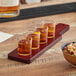 An Acopa mahogany flight paddle with three tasting glasses of brown liquid on a table with a bowl of mixed nuts.