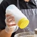 A person in gloves pouring sugar from a Vollrath Traex Polyethylene Shaker with a yellow lid into a bowl.
