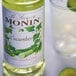 A close up of a Monin Premium Cucumber Flavoring Syrup bottle next to a glass of liquid.