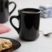 A white surface with two black Acopa Tiara stoneware mugs filled with coffee, a plate of scones, and cookies.