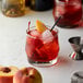 A glass of red liquid with ice and peach slices on a table with a bottle of Monin Premium Stone Fruit Flavoring Syrup.