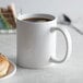 A white Acopa stoneware mug filled with brown liquid on a saucer with a donut.