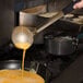 A person using a Vollrath Jacob's Pride ladle to pour sauce into a pot on a stove.