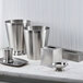 A brushed stainless steel pedestal wastebasket from the Focus Hospitality Pewter Veil Collection on a marble counter.
