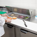 A chef in a white coat uses a knife with a black handle to prepare food on a Beverage-Air sandwich prep table.