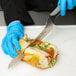 A person in blue gloves using a Dexter-Russell V-Lo Scalloped Offset Bread and Sandwich Knife to cut a sandwich.