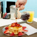 A person pouring Tablecraft syrup onto a stack of pancakes with strawberries.